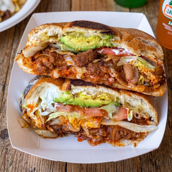 A large Mexican torta sandwich, cut in half, filled with meat, avocado, lettuce, and sauce.
