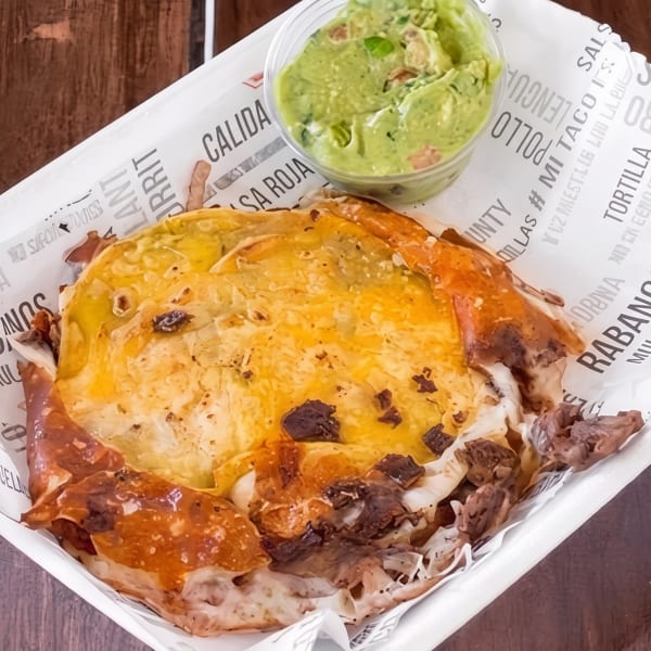 A cheesy quesadilla with meat and a side of guacamole in a white container on a wooden table.