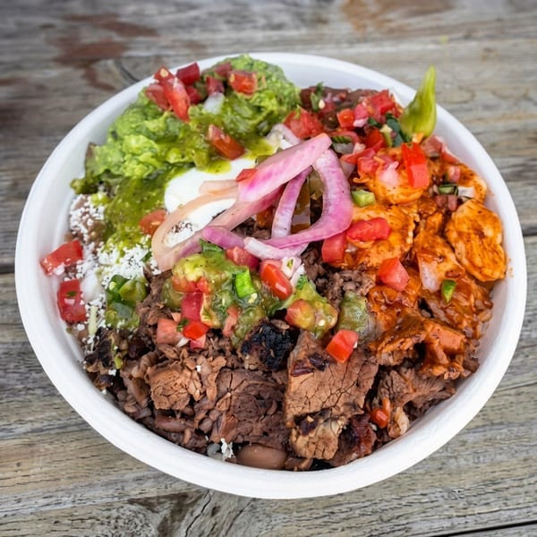 A vibrant burrito bowl with steak, shrimp, guacamole, salsa, pickled onions, and sour cream.