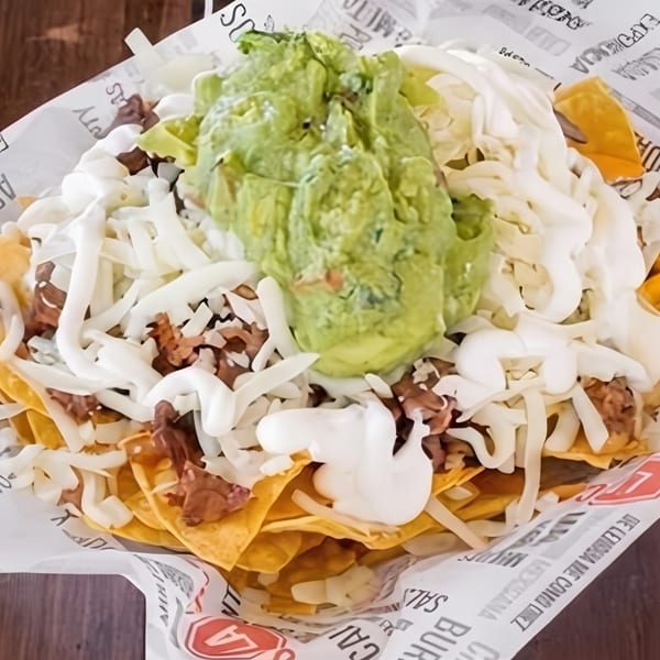 A close-up of loaded nachos with meat, shredded cheese, sour cream, and guacamole in a paper-lined basket.